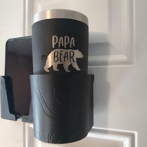 Può includere: Un bicchiere nero con bordo argentato e le parole "PAPA BEAR" e una silhouette di orso in bianco. Il bicchiere è in un supporto nero montato su una porta bianca.
