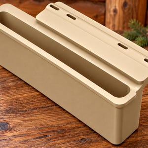 May include: A beige rectangular storage bin with a stepped top edge featuring three horizontal slots. The container has a deep, open compartment and a flat, extended mounting flange along the back side with three pre-cut holes for installation.