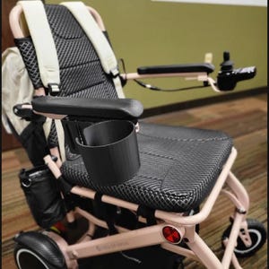 Puede incluir: Una silla de ruedas eléctrica de color rosa claro con asiento y respaldo de malla negra, correas de hombro color crema y un portavasos negro unido al reposabrazos. La silla incluye un control tipo joystick y ruedas negras.
