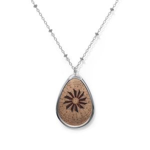 Puede incluir: Collar con colgante plateado con un colgante ovalado. El colgante presenta un fondo marrón con un diseño de estrella roja oscura y escritura manuscrita. La cadena del collar es de color plateado delicado.