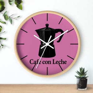 May include: A round wall clock with a light wood frame and a pink face. The center features a black graphic of a moka pot coffee maker. Below the graphic, the text reads Café con Leche in black. The clock has white hour, minute, and second hands.
