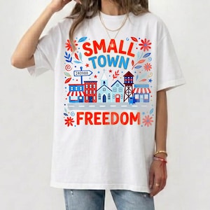 Könnte beinhalten: Weißes T-Shirt mit einem farbenfrohen Grafikdesign mit den Worten "SMALL TOWN FREEDOM" über einer Reihe stilisierter Gebäude. Das Design umfasst rote, blaue und grüne Blumen- und Sternakzente sowie ein Straßenschild mit der Nummer 240988.