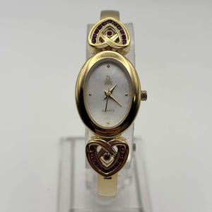 Op de afbeelding: Een goudkleurig quartzhorloge met een ovale wijzerplaat van wit parelmoer, gouden wijzers en indexen. De band bevat twee hartvormige schakels aan de boven- en onderkant, elk versierd met kleine rode steentjes in een goudkleurig metalen frame.