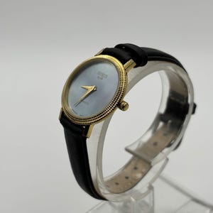 May include: A gold-toned wristwatch with a black leather band. The watch face is a light blue color with gold hands and the brand name "BRIEUX" printed on the face. The watch is displayed on a clear stand.