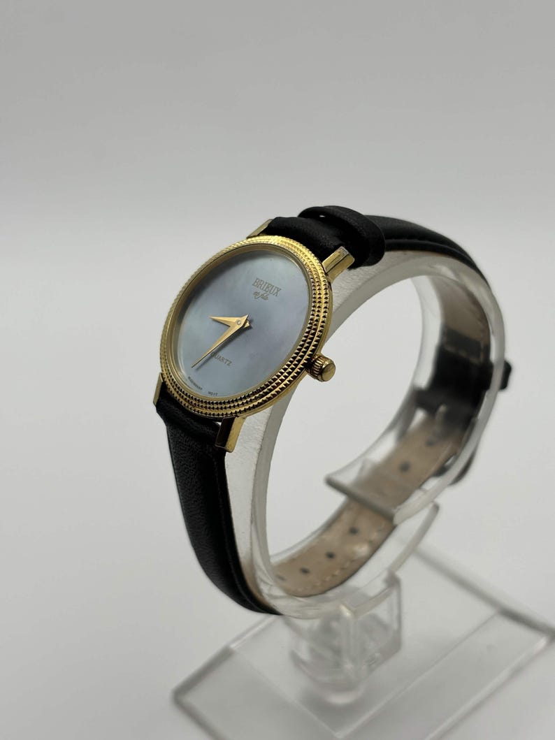 May include: A gold-toned wristwatch with a black leather band. The watch face is a light blue color with gold hands and the brand name "BRIEUX" printed on the face. The watch is displayed on a clear stand.