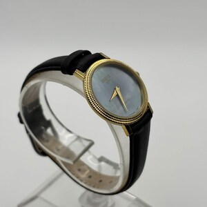 May include: A gold-toned wristwatch with a black leather band. The watch face is a light blue color with gold hands and a gold bezel. The watch is displayed on a clear stand.