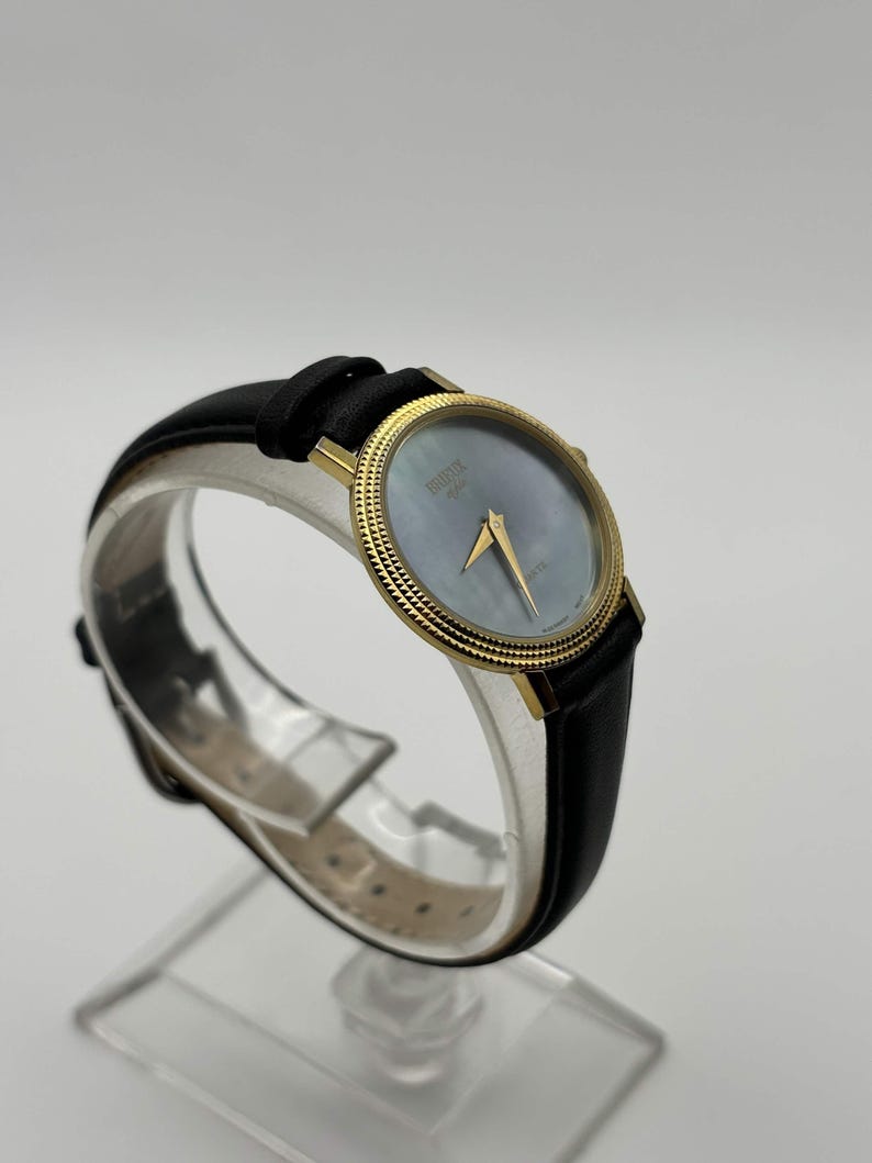 May include: A gold-toned wristwatch with a black leather band. The watch face is a light blue color with gold hands and a gold bezel. The watch is displayed on a clear stand.