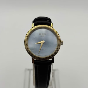 May include: A gold-toned wristwatch with a black leather band. The watch face is a light blue color with gold hands and the word "QUARTZ" printed on the face. The watch has the text "BRIEUX et fils" on the face.