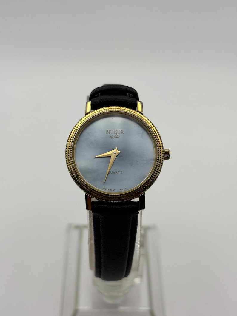 May include: A gold-toned wristwatch with a black leather band. The watch face is a light blue color with gold hands and the word "QUARTZ" printed on the face. The watch has the text "BRIEUX et fils" on the face.