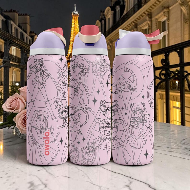 May include: Three pink Owala water bottles with black and white anime character illustrations. Each bottle has a light purple lid with a pink flip-top and a white handle. The background features a Parisian street scene.