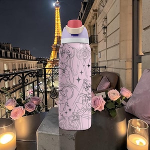 May include: A pink water bottle featuring a cartoon design with black outlines. The bottle has a light purple and red cap. The background shows the Eiffel Tower at night, with flowers and candles on a table.