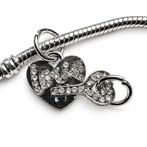 May include: A silver-toned snake chain bracelet featuring a heart-shaped charm with the word love spelled out in clear crystals and a separate key-shaped charm encrusted with clear crystals, both attached to the bracelet by a circular jump ring.