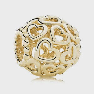 May include: A spherical gold charm bead featuring an intricate openwork design composed of multiple interconnected heart shapes. The surface of the bead displays a polished metallic finish with a repeating pattern of cut-out hearts throughout.