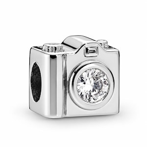 May include: A silver-toned charm shaped like a camera, featuring a polished metallic finish and a single, round, clear faceted stone set in the centre of the lens area. The charm includes a small top dial and a rectangular viewfinder detail.