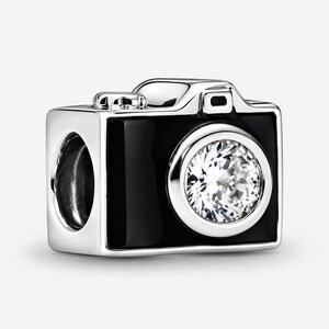 May include: A silver charm shaped like a camera with a glossy black enamel finish. The lens is represented by a large, round, clear faceted stone set in the centre. The top of the charm features a small silver button detail and a viewfinder.