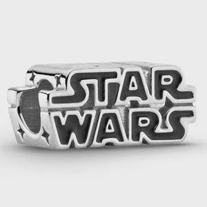 Op de afbeelding: Een zilverkleurige metalen bedel met het Star Wars-logo in zwarte emaille letters. Het rechthoekige ontwerp bevat kleine stervormige uitsparingen aan de zijkanten en een centrale horizontale opening voor een armband of ketting.