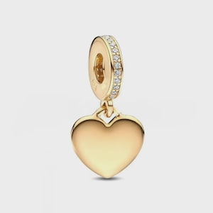 May include: A gold-toned heart-shaped charm pendant featuring a smooth, polished surface. The top bail is cylindrical and encrusted with a row of small, clear, sparkling stones along its outer edge, connecting to the heart via a small loop.
