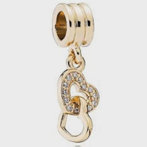 May include: A gold-toned charm bead featuring a cylindrical top section with three horizontal grooves. Suspended from this are two interlocking heart-shaped pendants, with the upper heart encrusted with small, clear, sparkling stones.