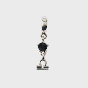 Può includere: Un ciondolo in metallo color argento con un occhiello circolare nella parte superiore, una singola pietra preziosa rotonda blu scuro sfaccettata incastonata in una montatura a griffe al centro e il simbolo zodiacale Bilancia in fondo.