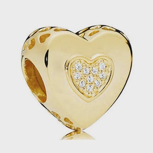 May include: A gold-toned heart-shaped charm bead featuring a smaller, raised heart centre encrusted with clear, sparkling stones. The sides of the bead are decorated with a repeating cutout heart pattern, creating a hollow, detailed metallic structure.
