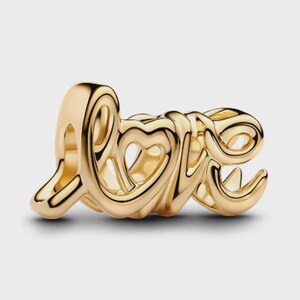 May include: A gold-toned metal charm shaped like the word love in a cursive script font. The design features a heart shape integrated into the letter o, with smooth, rounded edges and a polished finish that reflects light across the surface.