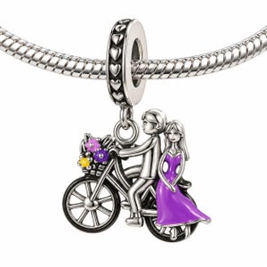 Può includere: Un ciondolo in argento a forma di bicicletta con un cestino di fiori viola e gialli, che trasporta due figure. Una figura è seduta sul sellino mentre l'altra è in piedi dietro con un abito viola. Il ciondolo ha un gancio con motivo a cuori.