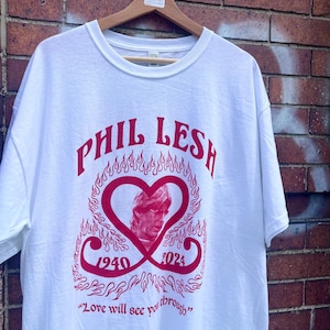 May include: White t-shirt with a red graphic featuring the text "PHIL LESH" above a heart-shaped design with a portrait and the years 1940-2024. The phrase "Love will see you through" is printed below the design.
