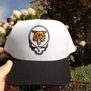 May include: A white and black baseball cap featuring an embroidered design. The design includes a skull with a snarling wildcat face in a circle. The cap has a black brim and a white crown.