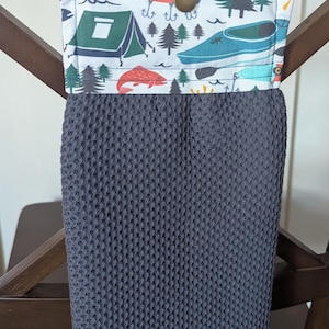 May include: A hanging kitchen towel featuring a dark gray waffle-textured fabric attached to a fabric topper. The topper has a white background with a colorful outdoor camping and fishing print, including tents, pine trees, fish, and lures, secured by a brass-colored snap button.