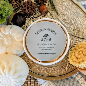 Könnte beinhalten: Ein festes Geschirrspülmittel von Hessler Hearth, mit dem Text "Fresh Mint and Eucalyptus". Die Seife befindet sich in einem weißen Behälter, neben zwei dekorativen Seifen und einer Holzspülbürste. Die Seifen haben ein Blumendesign.