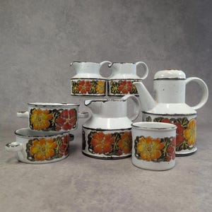 May include: A collection of white ceramic tableware with a floral design in orange, yellow, and red. The set includes a teapot, pitchers, small bowls, and a mug. The rims and handles are outlined in black.