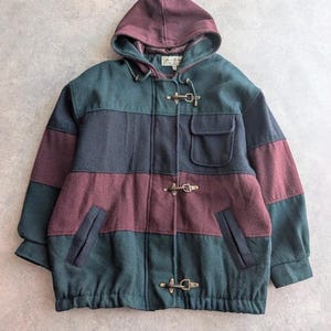 May include: A hooded jacket featuring a color-blocked design in dark green, black, and burgundy. The garment includes three metal toggle closures down the front, two side pockets, and one chest pocket with a flap. The fabric has a textured finish.