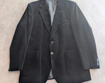 Pode incluir: Um blazer de veludo preto com lapela entalhada, dois botões frontais, dois bolsos grandes aplicados e quatro botões pequenos em cada punho. O casaco é totalmente forrado com um tecido cinzento escuro visível na gola.