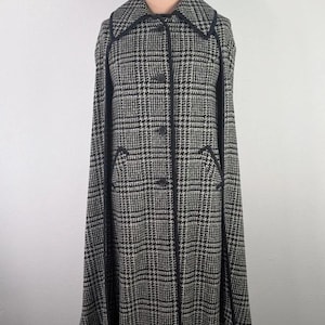 May include: A black and white glen plaid cape coat featuring a wide collar, a front button closure, and black trim along the edges. The garment has arm slits and a structured silhouette, displayed on a mannequin with the text IGD on the neck.