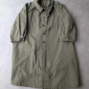 Puede incluir: Una gabardina de color verde oliva con cuello clásico, cierre frontal de botones con cuatro botones oscuros visibles y mangas largas con correas ajustables en los puños. El abrigo presenta un diseño de hombros estructurados.