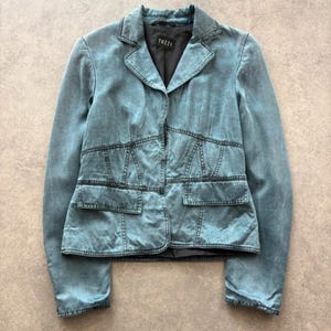 May include: A teal blue blazer with a washed denim appearance, featuring a notched lapel, long sleeves, two front flap pockets, and a black interior lining with a TUZZI brand label. The garment has visible dark stitching and structured seams.
