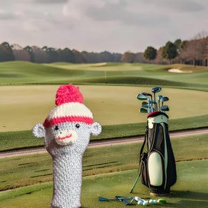 May include: A gray knitted golf club head cover shaped like a monkey with a red and white hat and a red pom-pom. A golf bag with clubs and golf balls are on a green golf course.