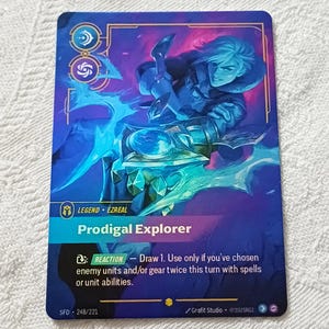 Peut inclure: Une carte à collectionner avec une illustration de personnage dans des tons bleus et violets. La carte indique "Prodigal Explorer" et comprend des instructions de jeu. La carte a une bordure bleue et le texte "LEGEND EZREAL" en haut.