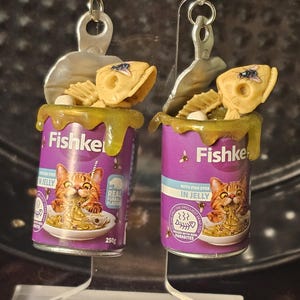 May include: Novelty earrings depicting miniature purple cat food cans labelled "Fishke". Each can has a cat illustration, a yellow topping, a fish skeleton, and a fly. The earrings hang from silver-coloured hooks.