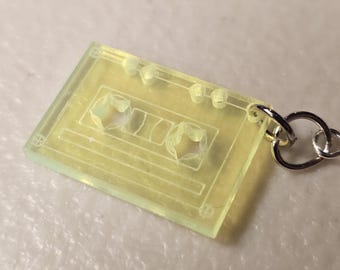 Puede incluir: Un colgante de casete amarillo translúcido con un llavero plateado. El colgante tiene forma rectangular con detalles grabados, incluyendo las bobinas de la cinta y otros elementos de diseño. El colgante está hecho de un material transparente.