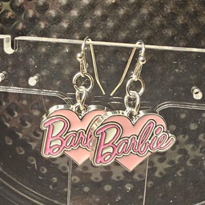 Può includere: Un paio di orecchini pendenti argentati. Ogni orecchino presenta un ciondolo a forma di cuore rosa con la scritta "Barbie" in corsivo, bordata di bianco e rosa. Gli orecchini sono sospesi a un gancio argentato.