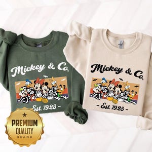 May include: Two crewneck sweatshirts, one olive green and one cream, each featuring a graphic of Disney characters in skeleton costumes. The text reads Mickey & Co. Est. 1928. The design includes a sunset-colored background with small bats flying above.