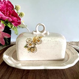 Peut inclure: Beurrier en céramique crème avec un motif de marguerites et de blé. Le plat rectangulaire a un couvercle assorti avec une poignée en anneau. Le plat repose sur un plateau festonné assorti. Un bouquet de fleurs roses est en arrière-plan.