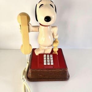 May include: A vintage Snoopy telephone in cream, holding a yellow handset. The phone has a red base with white buttons and a wooden-look base. A small yellow Woodstock figure is on the base.