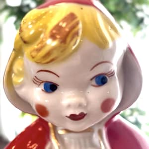 May include: A vintage ceramic figurine of a girl with blue eyes, rosy cheeks, and red lips. She wears a red hooded cape and a yellow cap with gold accents. The figurine has a glossy finish.