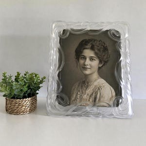 Peut inclure: Un cadre photo clair et orné avec un portrait de femme. Le cadre présente un motif décoratif tourbillonnant. Une petite plante en pot dans un panier tressé se trouve à gauche du cadre.