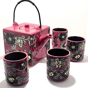 Peut inclure: Service à thé en céramique rose comprenant une théière carrée avec une anse noire et quatre tasses assorties. L'ensemble est orné d'un motif floral noir, blanc et gris, créant un contraste saisissant sur le fond rose.