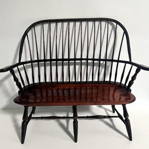 May include: A dark brown wooden Windsor-style bench featuring a high, arched back with vertical spindles, curved armrests, a smooth polished seat, and four tapered legs connected by a horizontal stretcher bar for structural support.