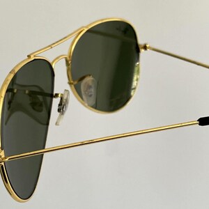 Puede incluir: Primer plano de gafas de sol aviador con montura dorada y lentes verde oscuro. Las gafas de sol tienen un doble puente y patillas negras. Las lentes son ligeramente reflectantes y la montura tiene un dise&ntilde;o cl&aacute;sico.