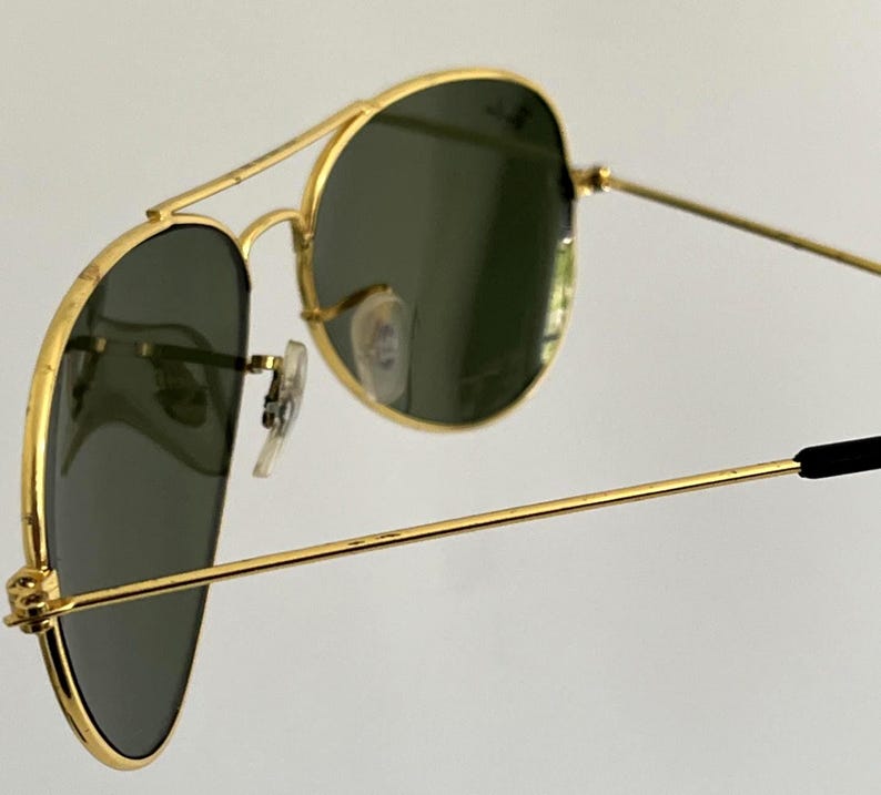 Puede incluir: Primer plano de gafas de sol aviador con montura dorada y lentes verde oscuro. Las gafas de sol tienen un doble puente y patillas negras. Las lentes son ligeramente reflectantes y la montura tiene un dise&ntilde;o cl&aacute;sico.